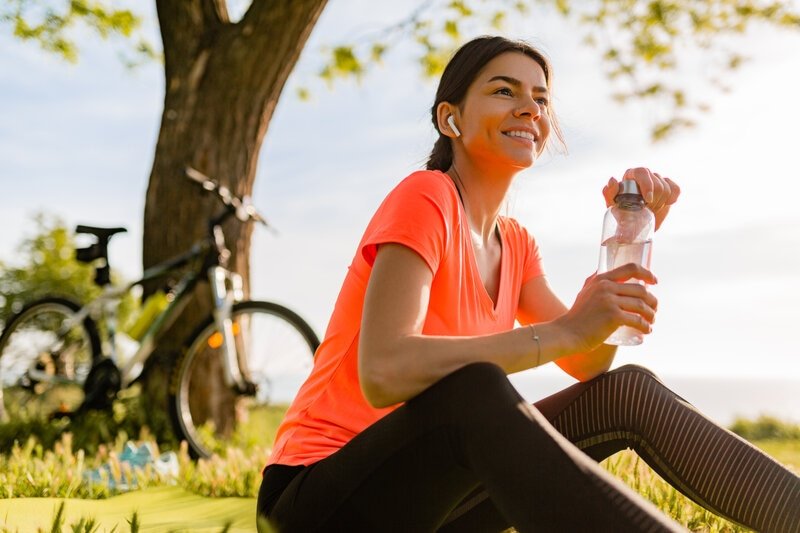 Mulher branca, sentada no parque, sorridente, com uma garrafa d'água na mão